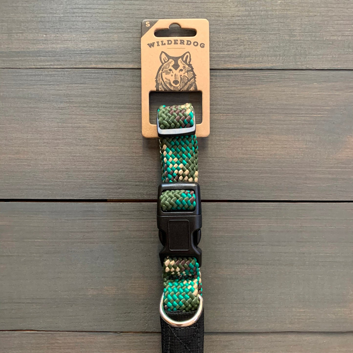 Displayed against a wooden background, the "Camo Collar" from Wilderdog introduces a vibrant and durable design in shades of green, blue, gray, and black. The packaging label at the top features the brand name "Wilderdog" along with an illustration of a dog's face. Crafted from rock climbing rope, this collar includes a black plastic buckle and metal ring for added strength and style.