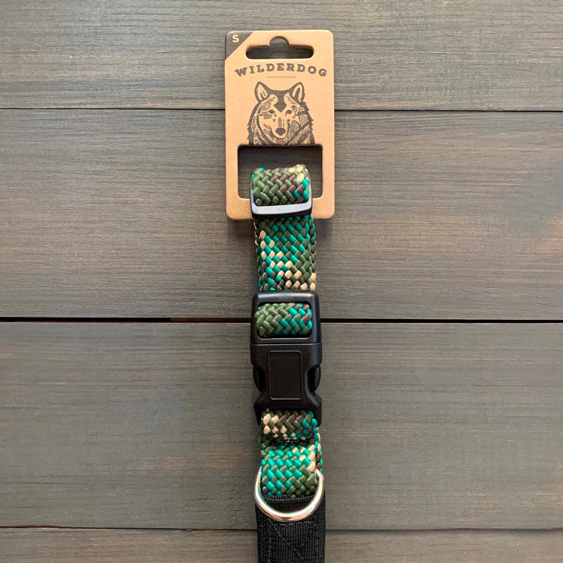 Displayed against a wooden background, the "Camo Collar" from Wilderdog introduces a vibrant and durable design in shades of green, blue, gray, and black. The packaging label at the top features the brand name "Wilderdog" along with an illustration of a dog's face. Crafted from rock climbing rope, this collar includes a black plastic buckle and metal ring for added strength and style.