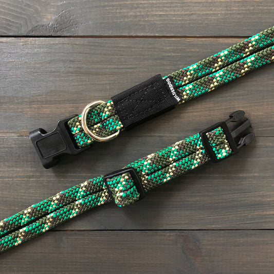 A close-up of two Camo Collars in green and black, featuring woven heavy-duty material with plastic buckle closures and metal D-rings, laid flat on a dark wooden surface. These collars have a textured pattern reminiscent of rock climbing rope and adjustable lengths with sliding buckles.
