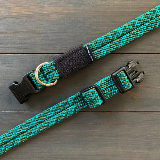 The Cascade Collar, a blue and green braided dog collar made from rugged rock climbing rope, includes a black plastic buckle and adjustment slider. Accompanied by a matching leash with a metal D-ring, it is showcased on a wooden surface.