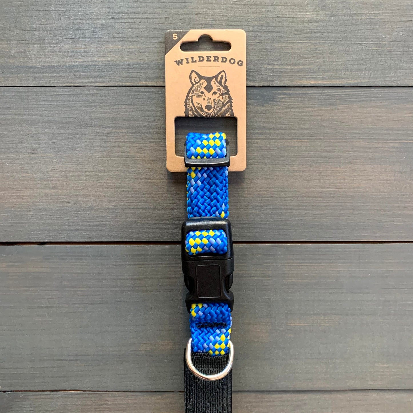 A durable Mariner Collar, adorned with yellow diamond patterns on a blue background, is displayed against a wooden surface. The collar features a black plastic buckle and a metal D-ring. The packaging label at the top reads "Wilderdog," showcasing an illustrated dog head alongside its promise of a lifetime warranty.