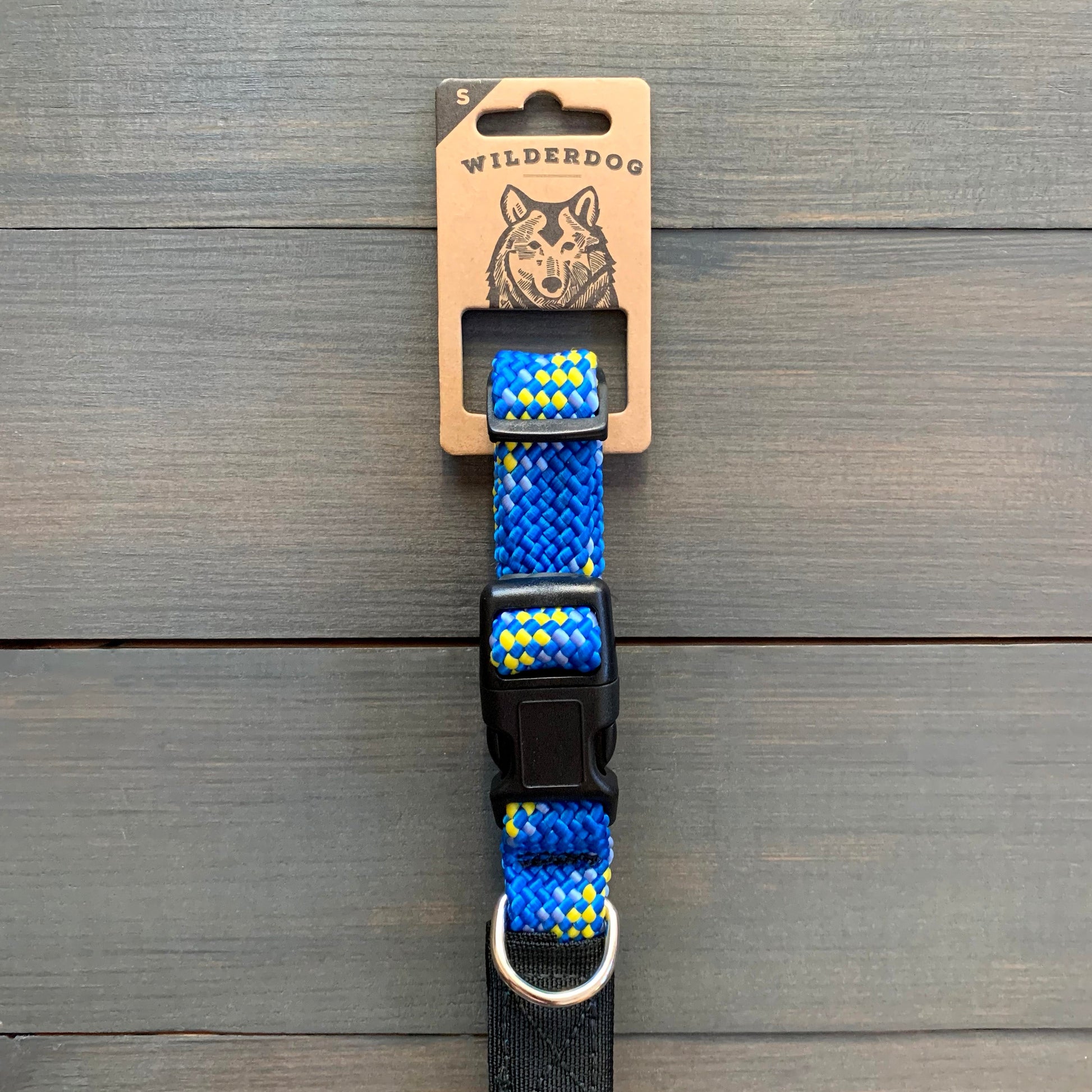 A durable Mariner Collar, adorned with yellow diamond patterns on a blue background, is displayed against a wooden surface. The collar features a black plastic buckle and a metal D-ring. The packaging label at the top reads "Wilderdog," showcasing an illustrated dog head alongside its promise of a lifetime warranty.