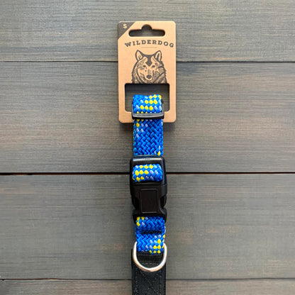 A durable Mariner Collar, adorned with yellow diamond patterns on a blue background, is displayed against a wooden surface. The collar features a black plastic buckle and a metal D-ring. The packaging label at the top reads "Wilderdog," showcasing an illustrated dog head alongside its promise of a lifetime warranty.
