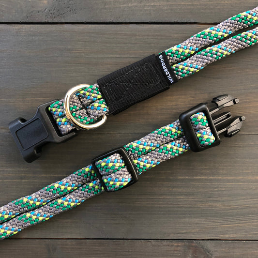 The Alpine Reflective Collars, a set of two durable dog collars crafted from braided rock climbing rope in green, blue, yellow, and gray hues, are displayed on a wooden surface. Each collar features black plastic buckles and metal D-rings for leash attachment.