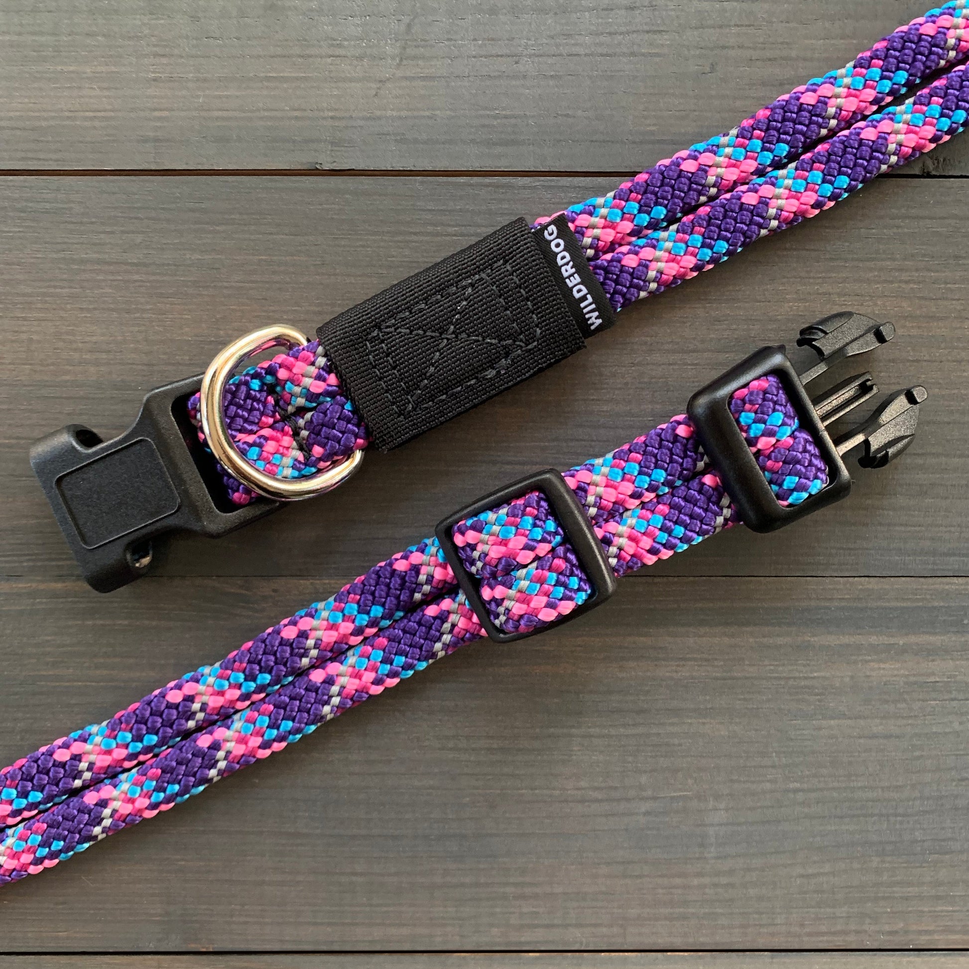 The Razzleberry Reflective Collar is a durable dog collar with a vibrant, multicolored design featuring shades of purple, pink, and blue. It includes a black plastic buckle, a metal D-ring for attaching a leash, and an adjustable strap. Covered by a lifetime warranty, the collar is laid out on a dark wooden surface.