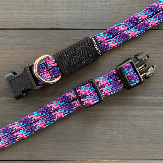 The Razzleberry Reflective Collar is a durable dog collar with a vibrant, multicolored design featuring shades of purple, pink, and blue. It includes a black plastic buckle, a metal D-ring for attaching a leash, and an adjustable strap. Covered by a lifetime warranty, the collar is laid out on a dark wooden surface.