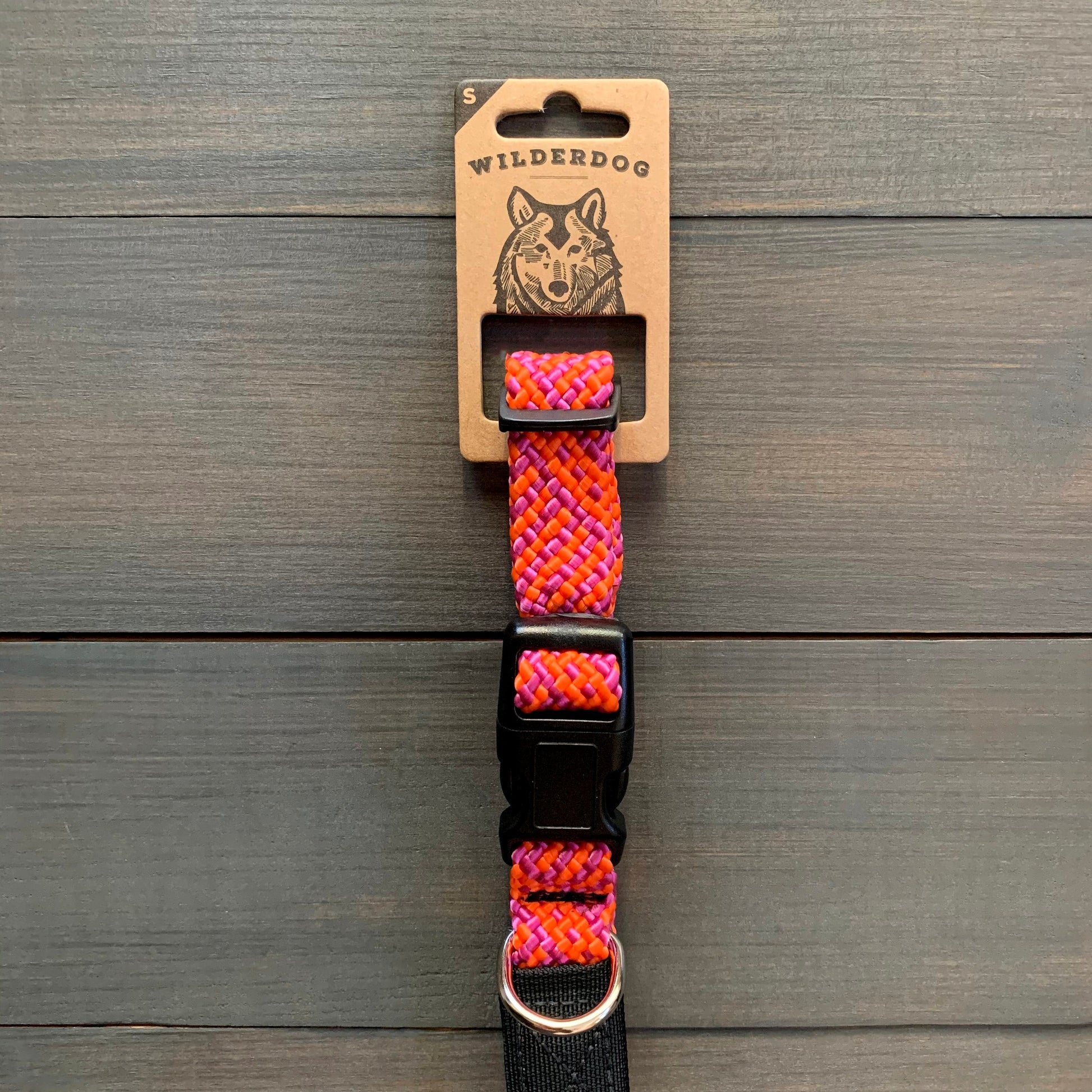 The Sierra Collar by Wilderdog, designed for small dogs, is displayed against a wooden background. This durable collar showcases a vibrant, woven pattern in shades of pink, orange, and purple. It features sturdy black hardware including a plastic buckle and a metal D-ring for leash attachment.