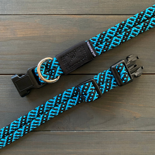 The Teton Collar, a stylish black and blue rock climbing rope collar, features a plastic side-release buckle and a metal D-ring for attaching a leash. Displayed on a wooden surface, the collar showcases a woven pattern and is padded for added comfort, ensuring durability and comfort for your dog.