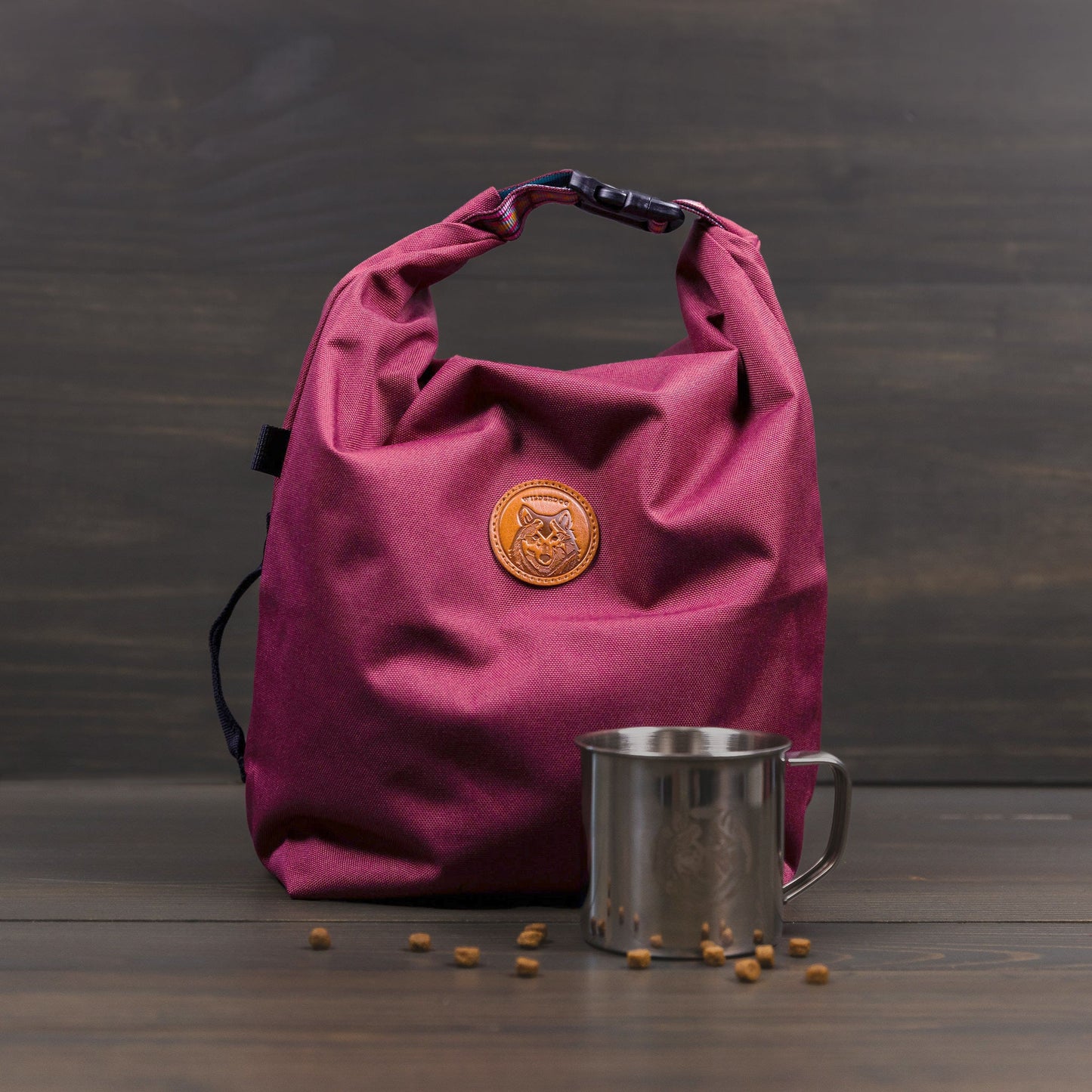 A Doggie Bag in maroon fabric with a rolled top and a logo-stamped leather patch sits on a wooden surface, ideal as a kibble carrier. Next to it, a stainless steel mug shines, surrounded by scattered small brown seeds against a backdrop of dark wooden planks.