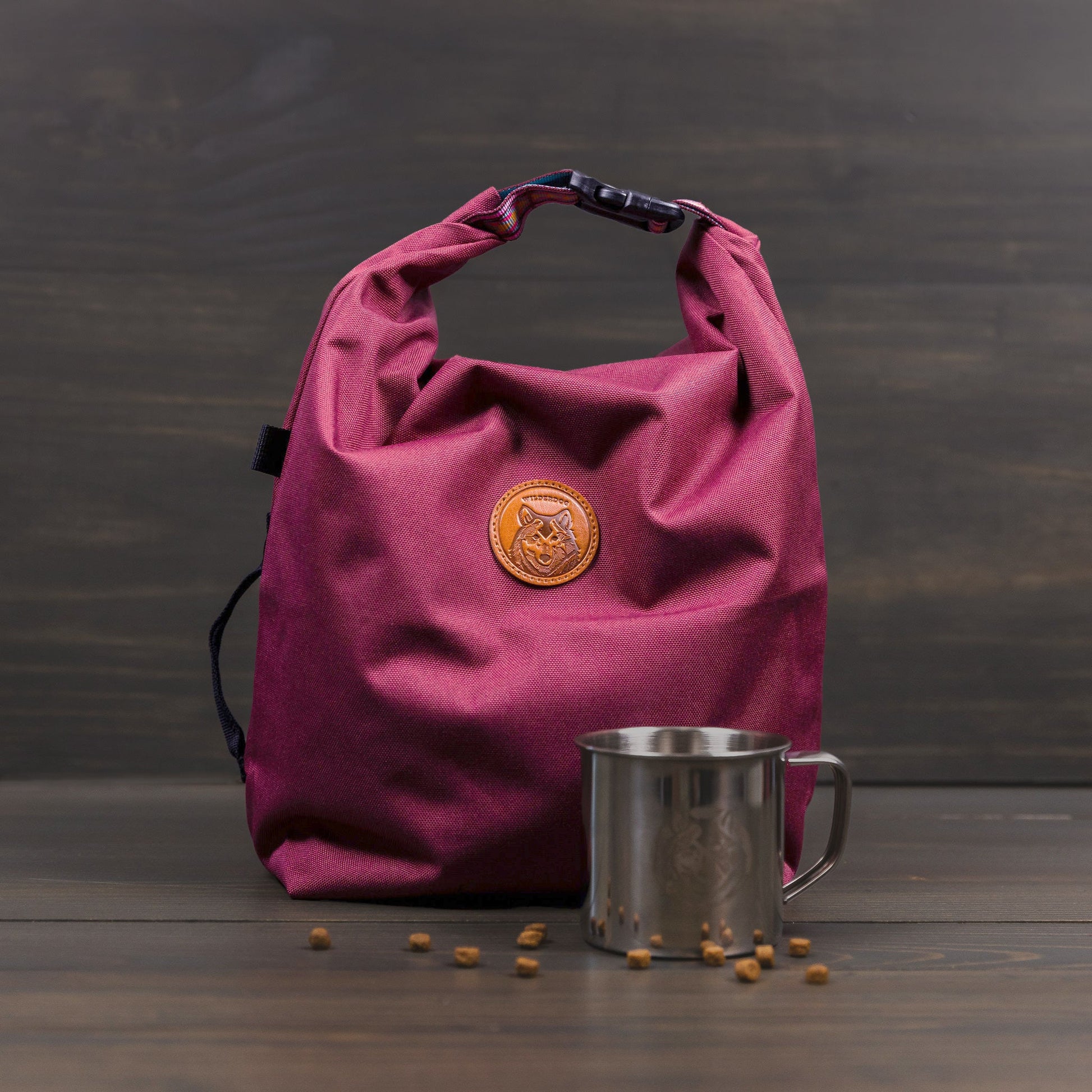 A Doggie Bag in maroon fabric with a rolled top and a logo-stamped leather patch sits on a wooden surface, ideal as a kibble carrier. Next to it, a stainless steel mug shines, surrounded by scattered small brown seeds against a backdrop of dark wooden planks.