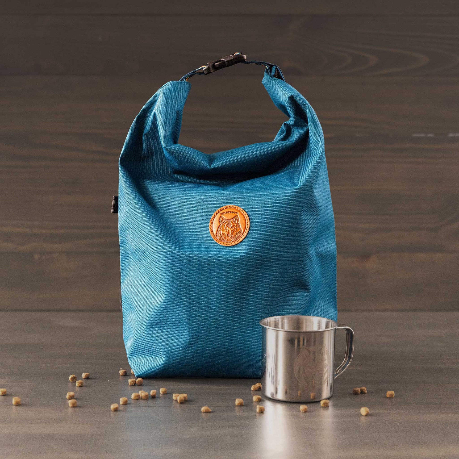 A blue roll-top Doggie Bag with an orange patch sits on wood as the ideal dog food travel bag, next to a stainless steel mug. Small pebbles scatter on the surface, set against a dark wooden wall backdrop.