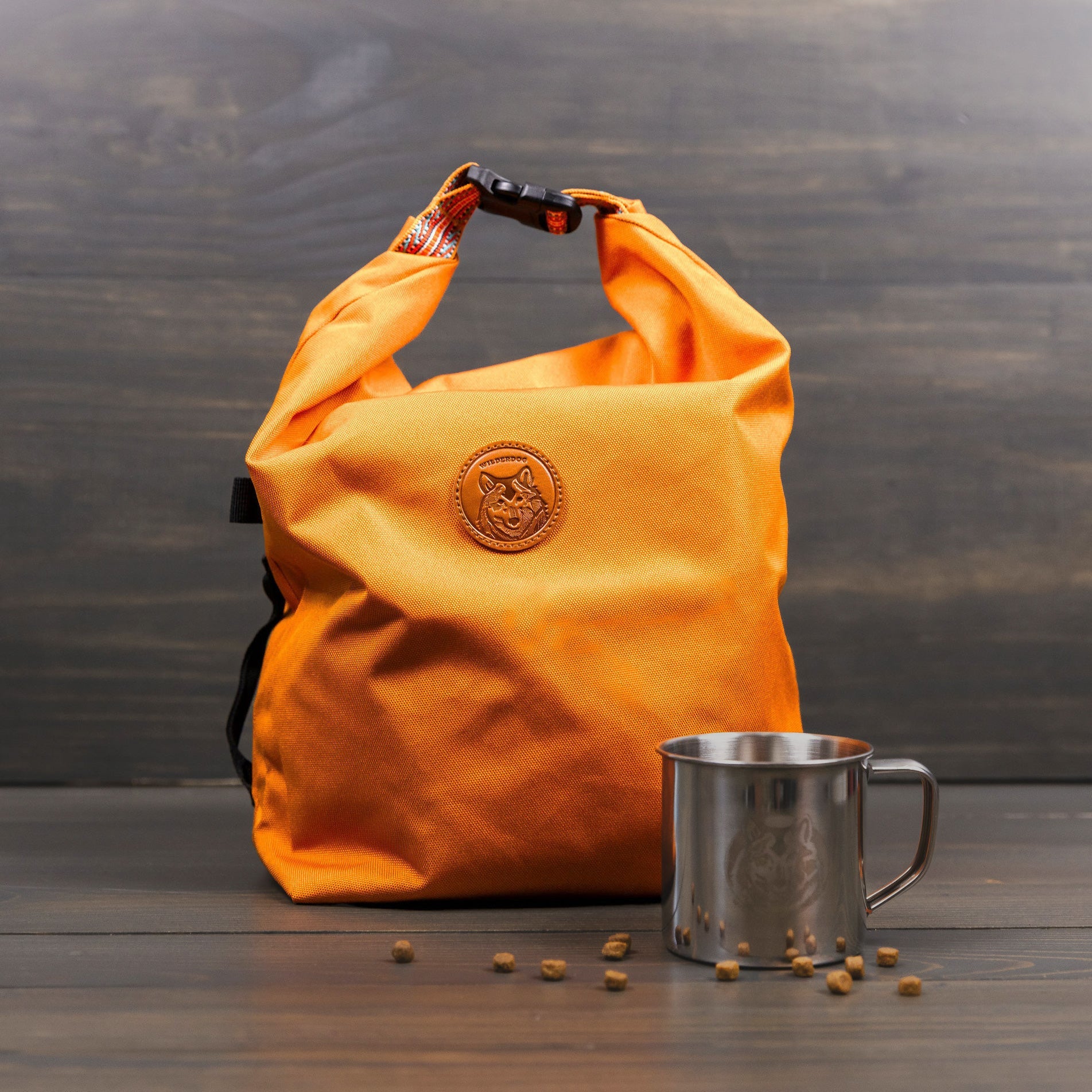 A vibrant orange Doggie Bag with a circular logo rests on a wooden surface beside a stainless steel mug, perfect for backpacking. Small brown seeds are scattered in front of the mug, set against the backdrop of a dark, wood-textured panel.