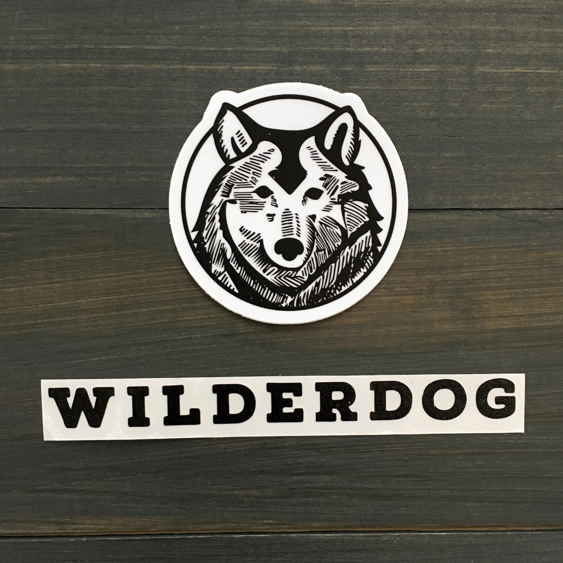 A black and white waterproof vinyl decal featuring a detailed illustration of a wolf's head inside a circle is placed on a dark wooden surface. Below the Wilderdog Sticker Decals, the word "WILDERDOG" is written in bold, black, uppercase letters.