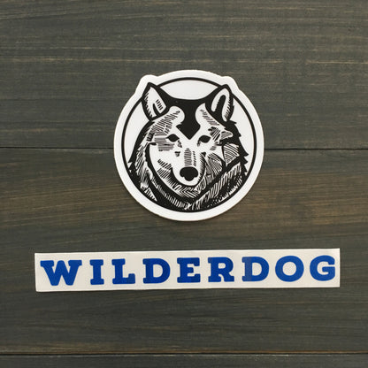 A circular logo showcases a black and white graphic illustration of a wolf's head. Below the logo, "WILDERDOG" is prominently displayed in bold blue capital letters. The background is a dark wooden surface, ideal for highlighting the durability of these outdoor-rated Wilderdog Sticker Decals.
