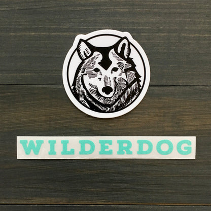 A black and white Wilderdog Sticker Decal showcases a stylized wolf's head inside a circle. Below the decal, the word "WILDERDOG" appears in bold turquoise uppercase letters. These outdoor-rated decals are set against a background of dark, wooden planks.