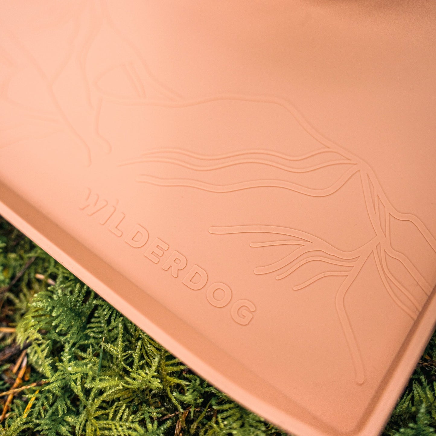 A close-up of a tan Silicone Placemat features an embossed mountain design and "WILDERDOG." It rests on green mossy ground, enhancing its natural, outdoors feel.