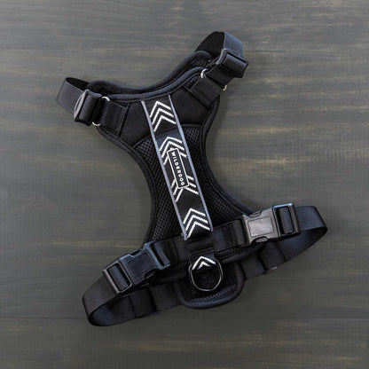 A Dog Harness in black, featuring adjustable straps and buckle closures, is laid flat on a dark wooden surface. Perfect for outdoor adventures, it boasts a pattern of white chevrons down the center, made with reflective fabric for enhanced visibility, and includes a metal ring for leash attachment.