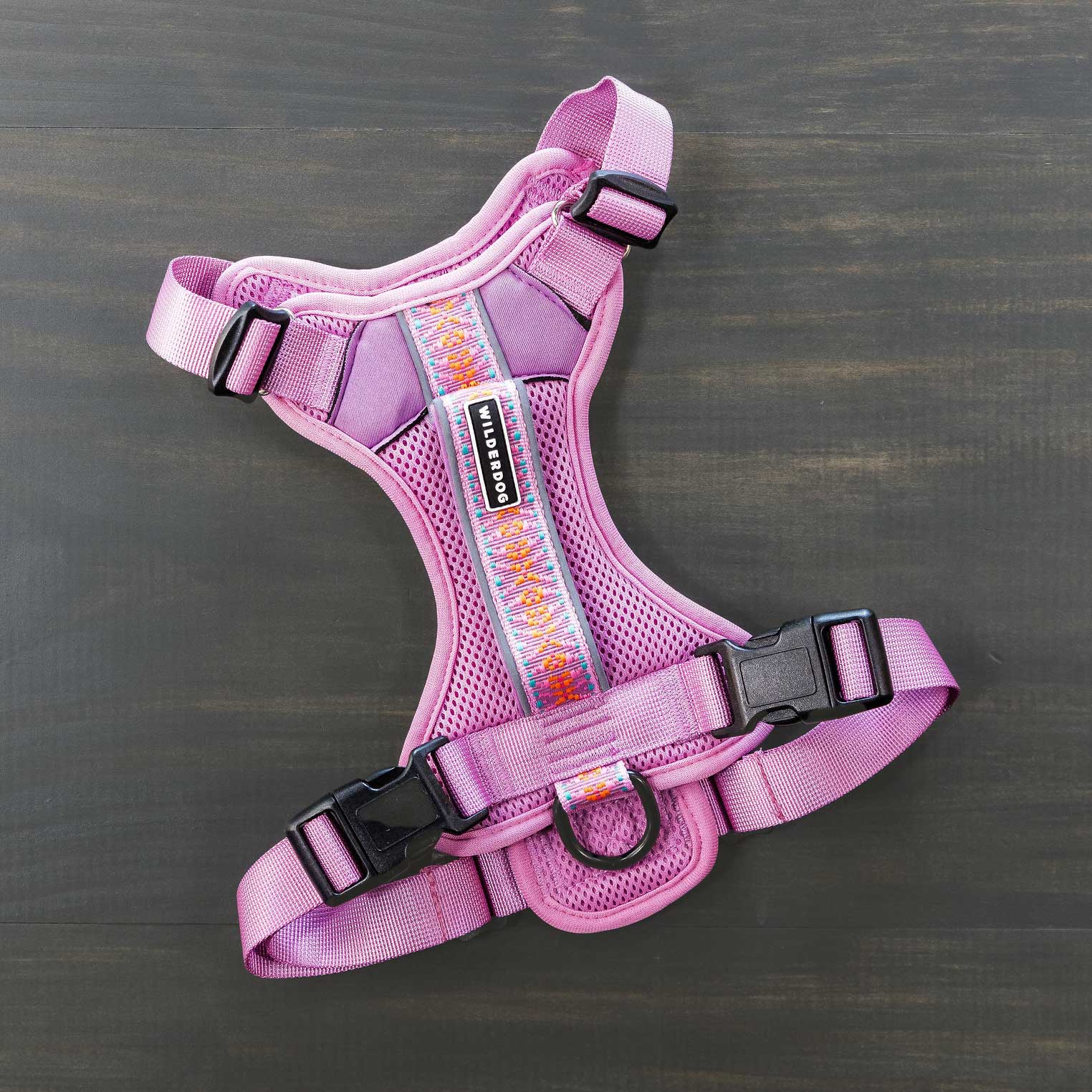 A Dog Harness, characterized by its lightweight pink design with black buckles and an adjustable strap, showcases a rainbow-patterned stripe down the center as it rests on a dark wooden surface. Its durable mesh construction guarantees comfort, and the reflective fabric boosts visibility for evening walks.