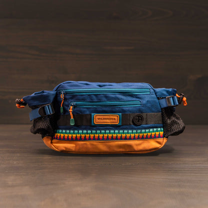 Introducing the Mega Hip Pack, featuring a vibrant blue top and an eye-catching orange base. It includes black side mesh pockets for added utility, a front zip pocket, and stylish patterned trim. The pack is complemented by an adjustable strap and showcased against a wooden background.