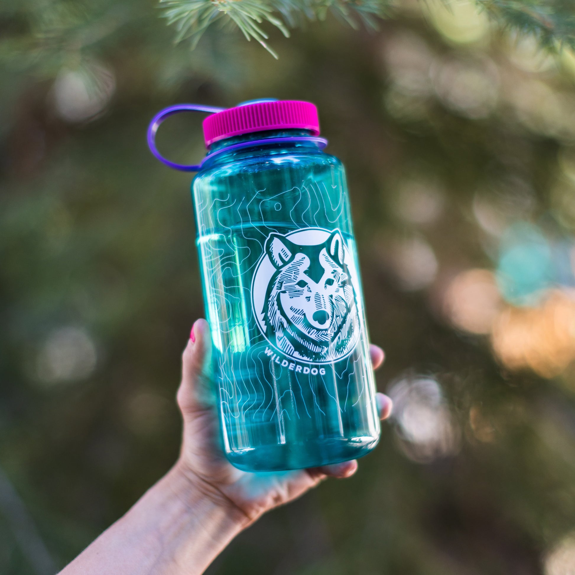 A hand holds a teal Wilderdog Nalgene water bottle with a purple cap and a white wolf logo on it. The 32oz Wide Mouth bottle, BPA/BPS-free for your safety, is set against a blurred natural background of greenery, hinting at an outdoor adventure.