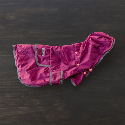 The Dog Rain Jacket, featuring a maroon and gray waterproof design with a hood and snap buttons, is laid flat on a dark surface. It includes a breathable mesh interior, reflective trim, a small brand label on the pocket, and velcro straps for securing.