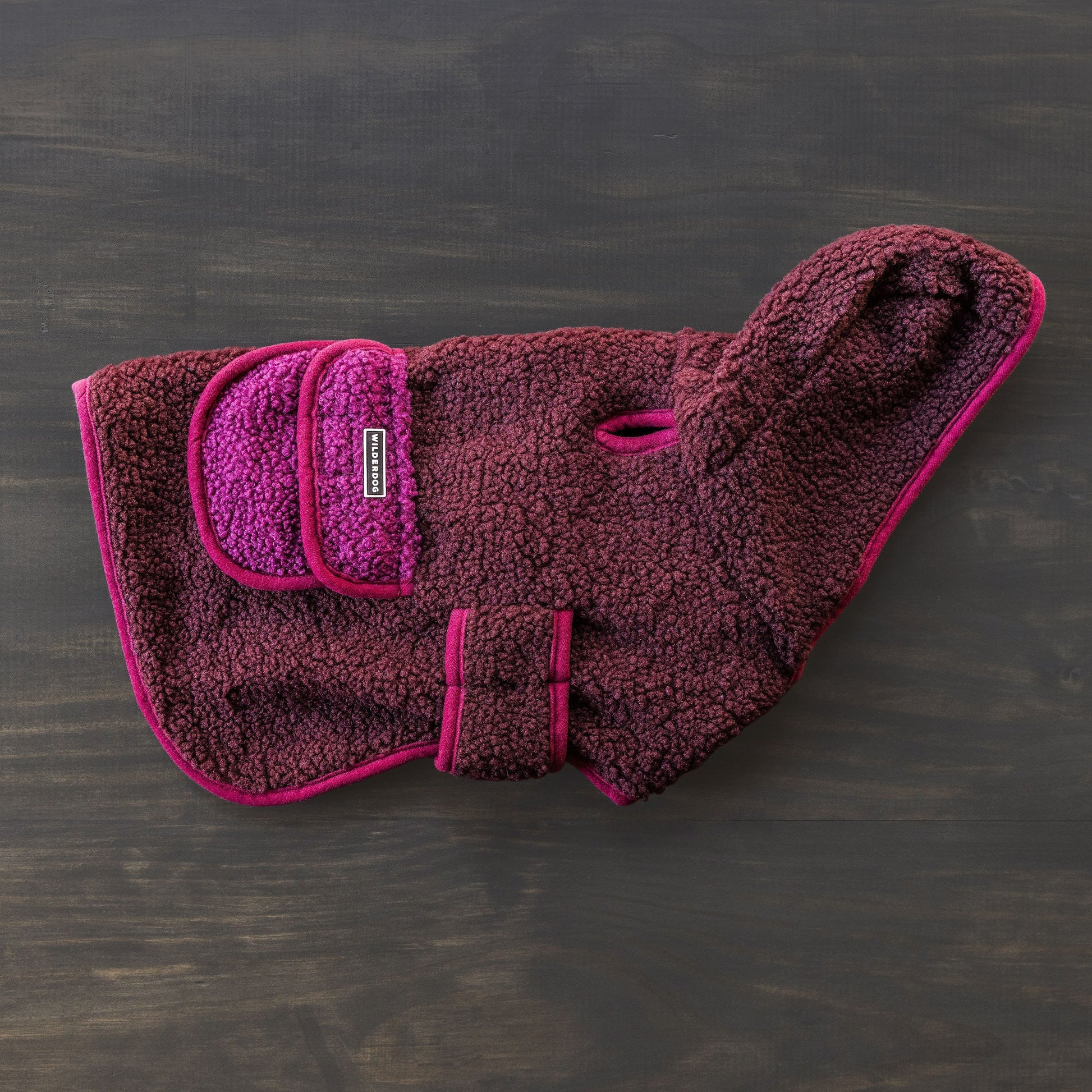 A Sherpa Jacket in pink and purple, crafted from soft, textured fleece fabric, is displayed against a dark background. This dog coat includes a pocket, an adjustable strap, and a handy leash hole for extra warmth and comfort.