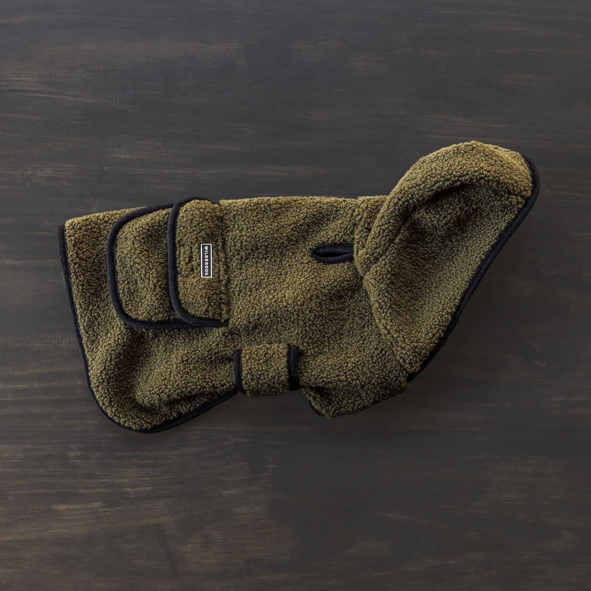 The Sherpa Jacket, a green fleece dog coat with black trim and adjustable straps, is displayed on a dark wooden surface. This cozy jacket includes a small leash hole for added convenience.