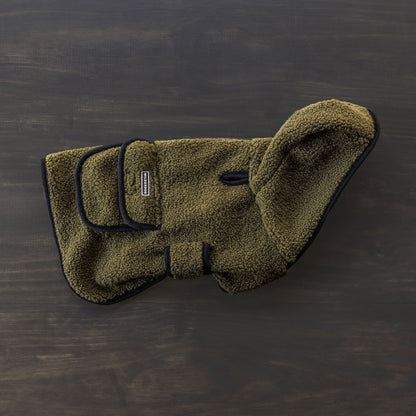 The Sherpa Jacket, a green fleece dog coat with black trim and adjustable straps, is displayed on a dark wooden surface. This cozy jacket includes a small leash hole for added convenience.