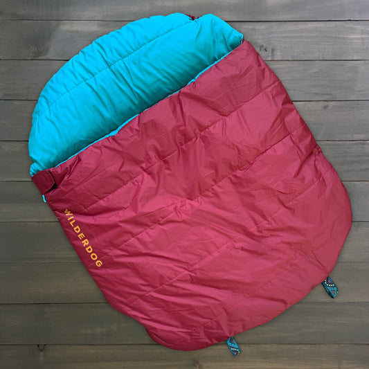Red and blue "Sleeping Bag" with "WILDERDOG" branding in yellow, laid on a wooden floor.