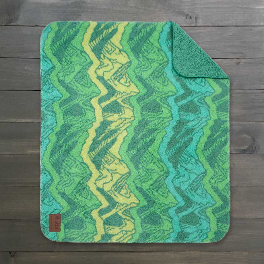 The Sherpa Fleece Waterproof Blanket, featuring a vibrant green and yellow zigzag pattern on a wooden background, showcases its cozy texture. The top right corner is slightly folded to reveal its plush feel, with a small brown tag in the lower left.