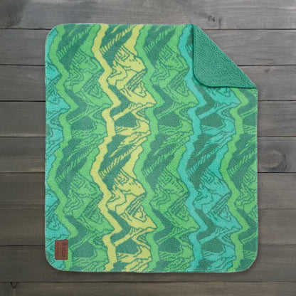 The Sherpa Fleece Waterproof Blanket, featuring a vibrant green and yellow zigzag pattern on a wooden background, showcases its cozy texture. The top right corner is slightly folded to reveal its plush feel, with a small brown tag in the lower left.