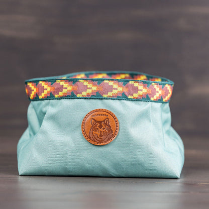 A light blue fabric bag featuring a colorful, patterned strap at the top and a front round leather patch embossed with an animal design, possibly a wolf. Ideal for outdoor adventures, it pairs perfectly with the Backpacking Bowl to cater to your furry companion's needs.