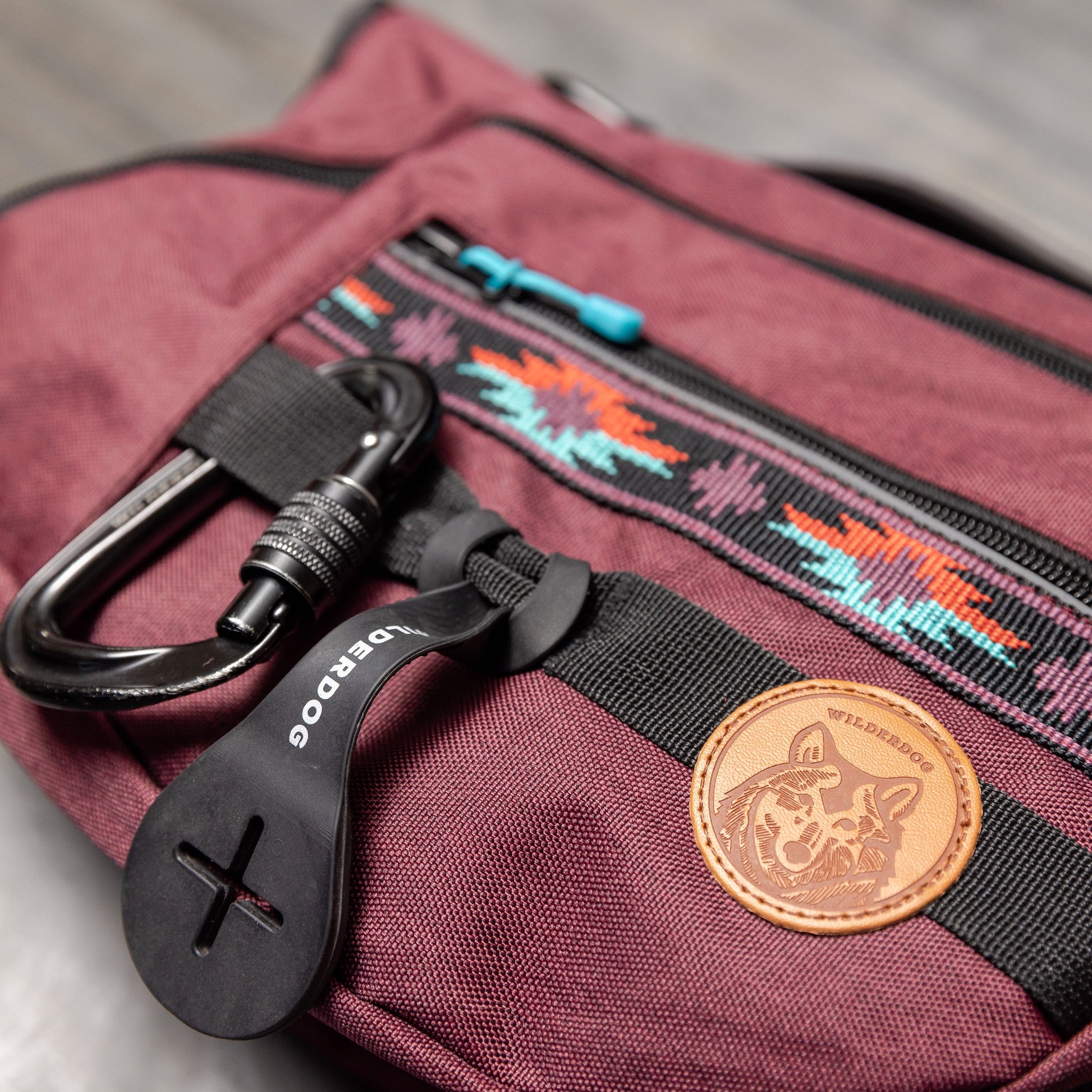A close-up photo of the Dog Backpack in burgundy, featuring black zippers and a front pocket adorned with a colorful Native American-inspired pattern. Attached to the pocket is a black carabiner clip on a black plastic tag, complemented by a brown leather patch showcasing a dog logo.