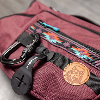 A close-up photo of the Dog Backpack in burgundy, featuring black zippers and a front pocket adorned with a colorful Native American-inspired pattern. Attached to the pocket is a black carabiner clip on a black plastic tag, complemented by a brown leather patch showcasing a dog logo.