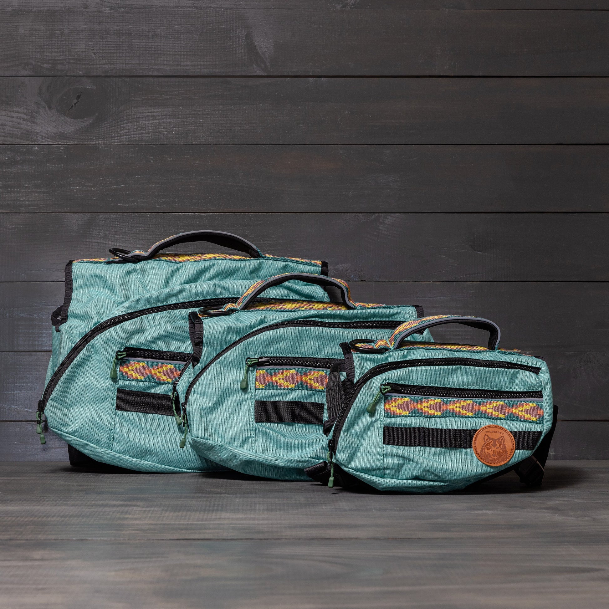 Three turquoise Dog Backpacks of different sizes are arranged on a dark wooden floor against a matching wooden wall. Each backpack, ideal as a hiking accessory, features a multicolored woven trim and a small brown leather patch with a logo. The backpacks have black zippers and handles.
