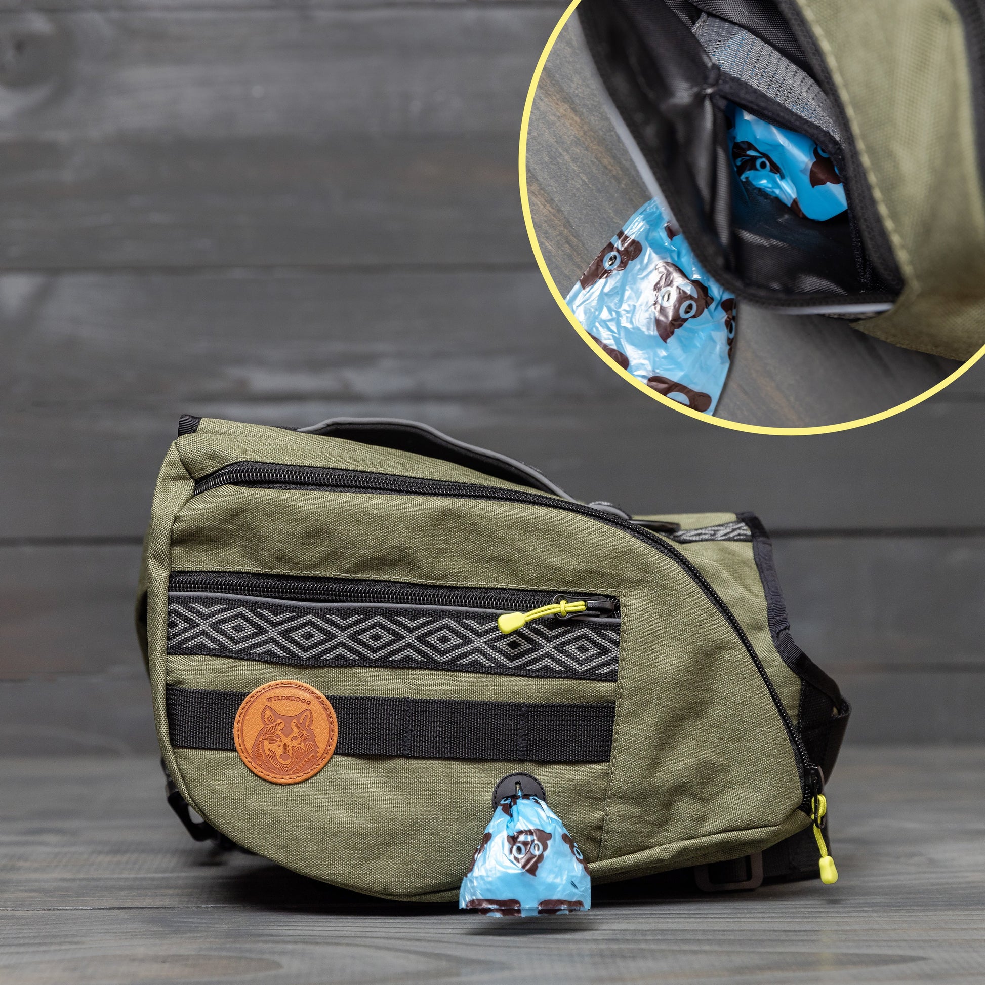 A khaki green Dog Backpack featuring black zippers and a decorative patch is displayed. A close-up inset reveals an outer pocket holding a small blue plastic bag adorned with a black cat pattern. Ideal as a hiking accessory, the backpack is set against a wood background.