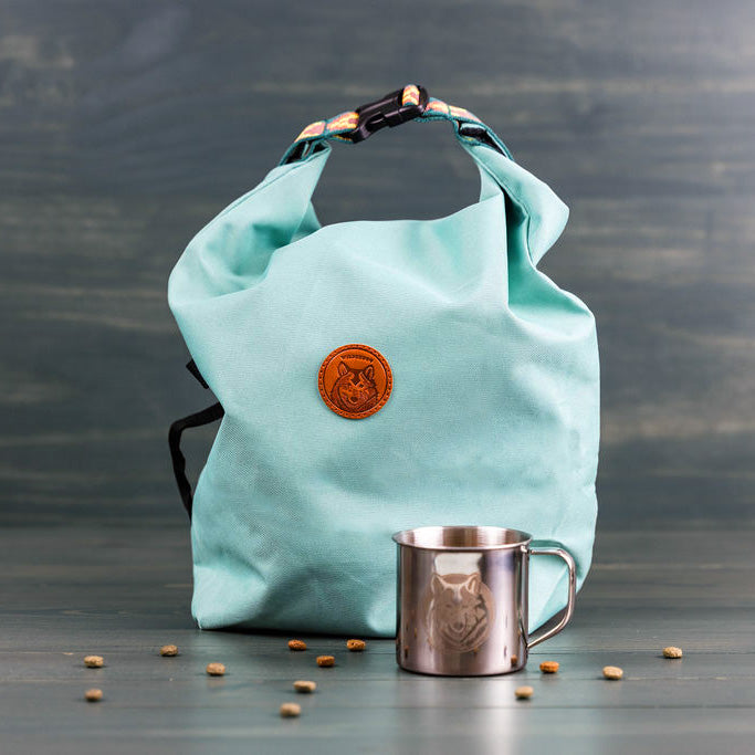 A light blue Doggie Bag roll-top backpack with a circular brown leather patch featuring an emblem in the center stands on a wooden surface. Beside it is a stainless steel mug etched with a wolf graphic, while small scattered items, possibly from the portable kibble carrier or dog food storage, are on the surface.