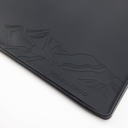 A detailed close-up of the Silicone Placemat reveals its black, rectangular form with a mountain design embossed in the top left corner. The word "WILDERDOG" is prominently embossed at the bottom center of this food-grade silicone mat. The background is white.