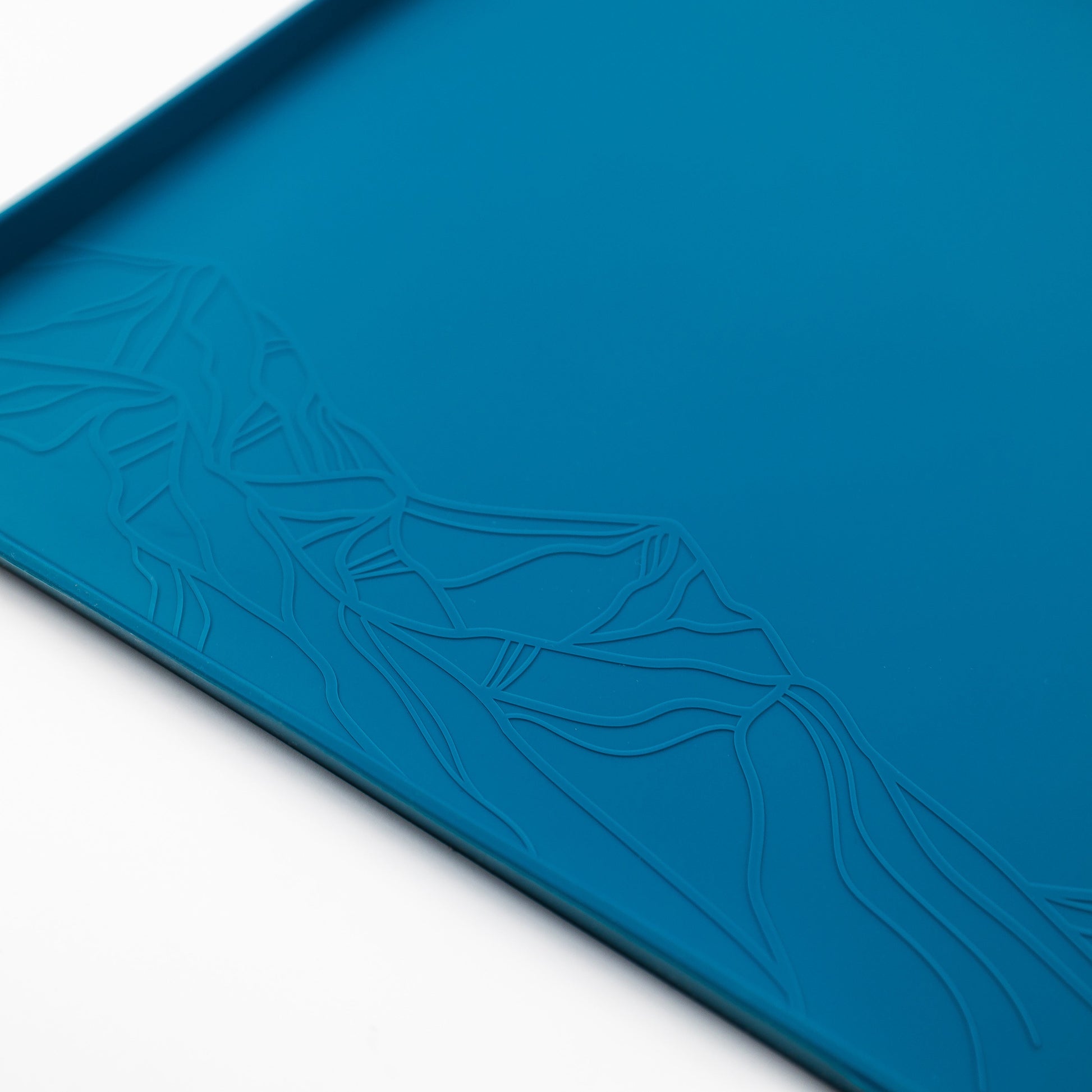Close-up of the Silicone Placemat, featuring a rectangular, blue design made from food-grade silicone. The mat showcases an embossed pattern depicting a stylized mountain range. Set against a white background, the intricate design is highlighted in one corner of the non-slip placemat.