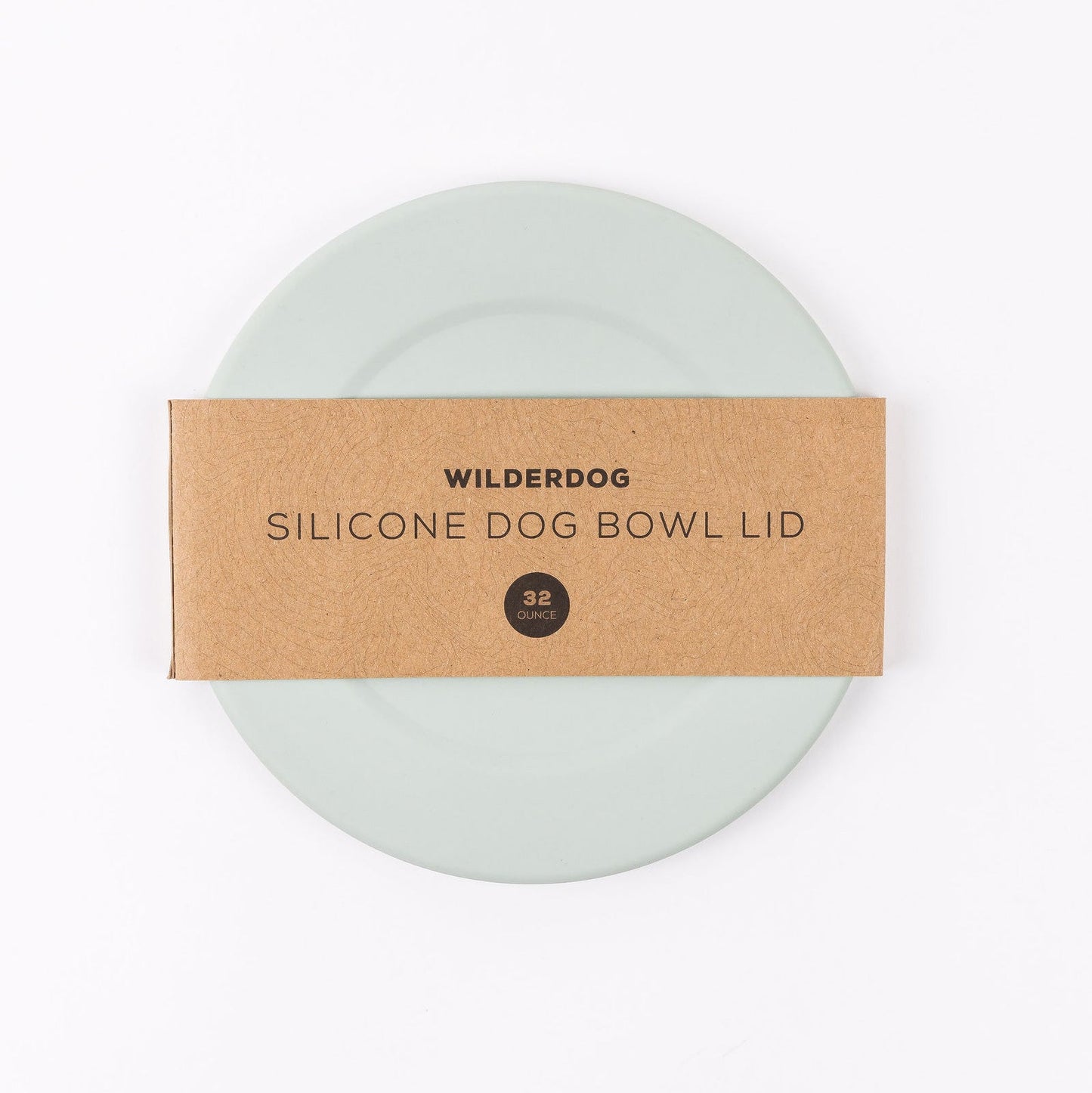 A round, light green silicone bowl lid made from food-grade silicone is partly covered by a brown paper band labeled "Wilderdog Silicone Dog Bowl Lid 32 Ounce." The lid, which is dishwasher safe, is placed against a plain white background.