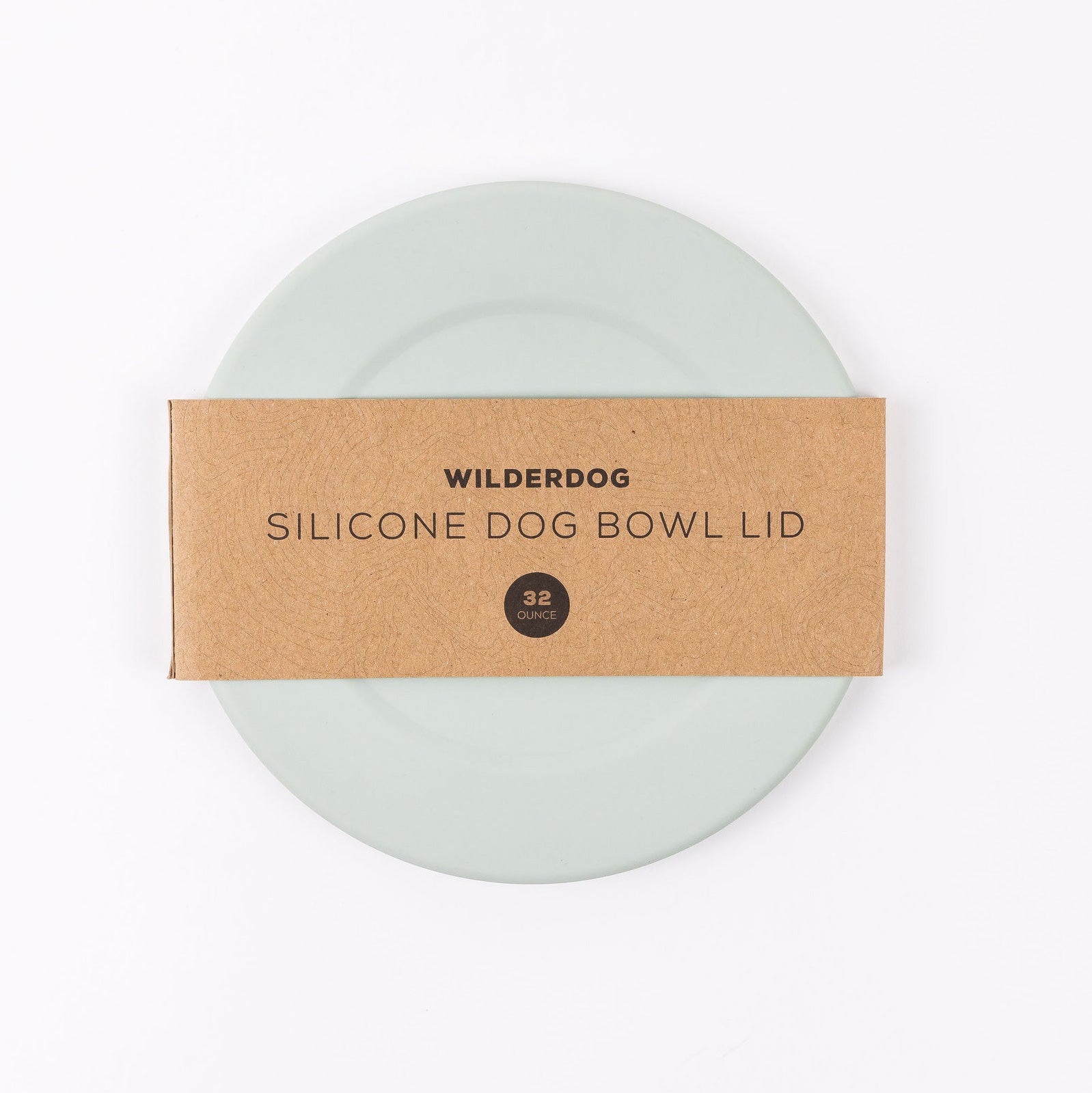 A round, light green silicone bowl lid made from food-grade silicone is partly covered by a brown paper band labeled "Wilderdog Silicone Dog Bowl Lid 32 Ounce." The lid, which is dishwasher safe, is placed against a plain white background.