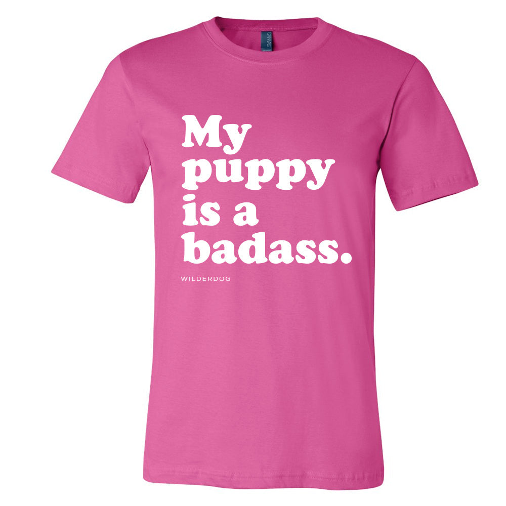 The Party Dog Tee Shirt is a pink, unisex t-shirt featuring bold white text on the front that says, "My puppy is a badass." The brand name "WILDRDOG" is printed in smaller white letters below the main text. It boasts vibrant colors and is made with sustainably produced materials.