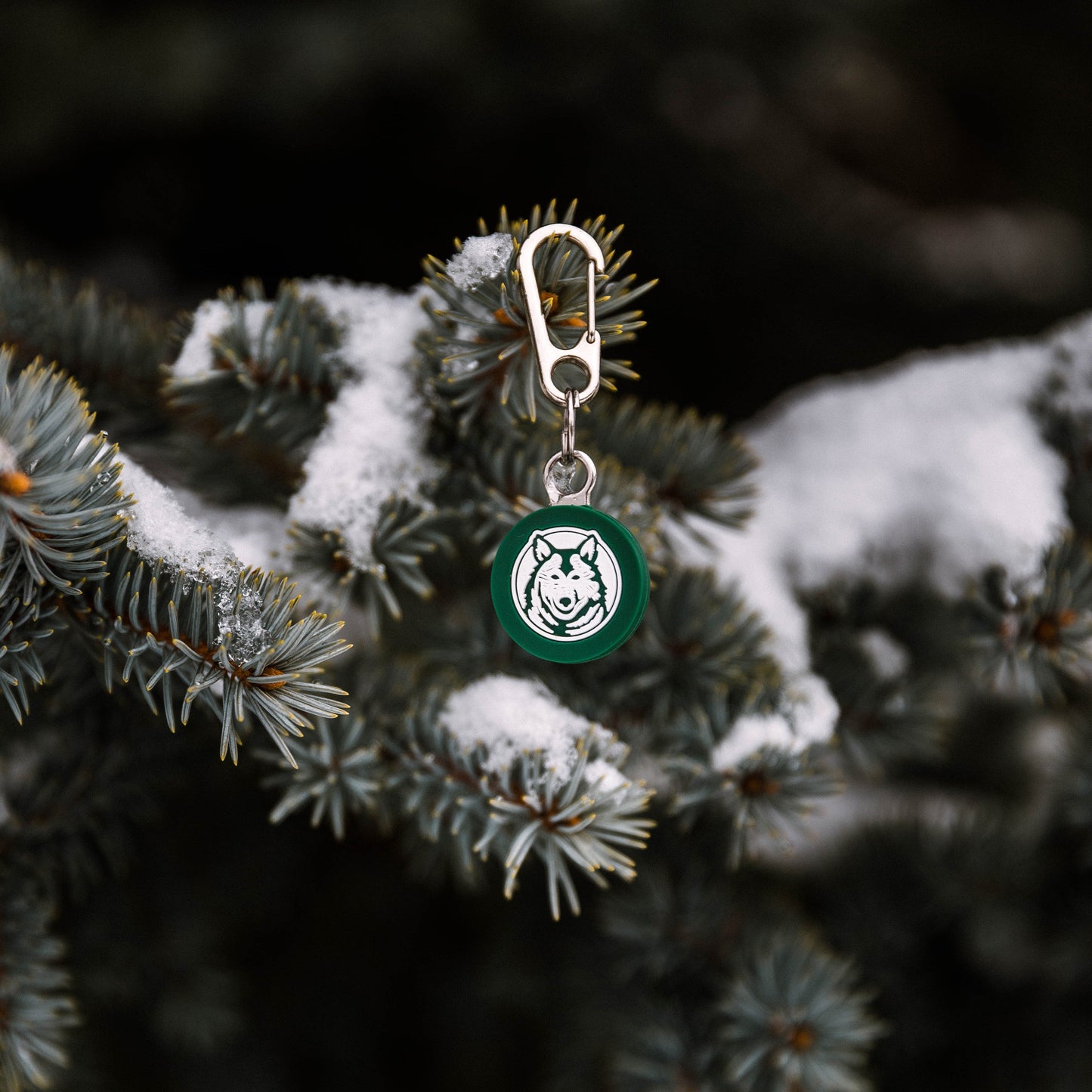 A Wilderdog Silicone Tag with a green circular charm, featuring a white abstract wolf design, dangles from a snow-covered evergreen branch, secured by a mini carabiner. The metal clasp of the keychain is attached to the charm. The background is a blurred view of more snowy branches.
