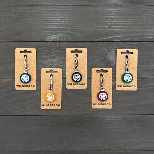Five Wilderdog Silicone Tags in various colors with mini carabiners on a dark wooden surface.